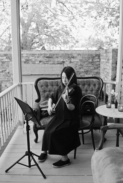 Wedding violinist performing live wedding music on a brick-walled porch, reading from a music stand with trees softening the background.