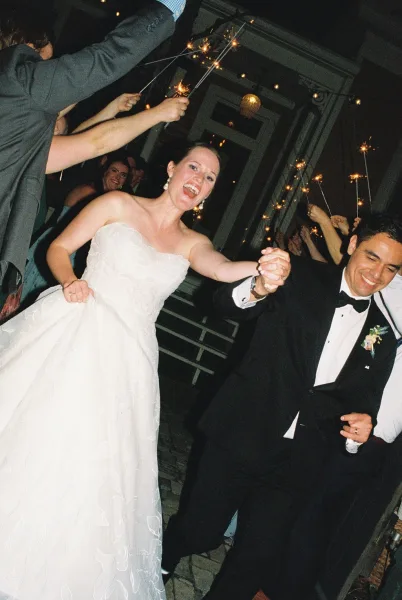 Sparkler exit with a wedding couple celebrating as they walk down the steps, guests cheering under string lights, groom in a black bow tie.