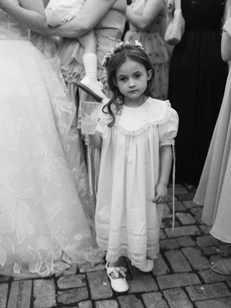 Flower girl portrait offering flower girl dress inspiration as she stands in a white smocked dress with a flower crown amid wedding guests.