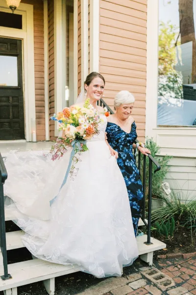 Bride with mother in a tender mother of the bride photo, arm in arm on the house porch steps as she holds a bouquet tied with a ribbon.