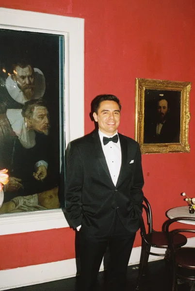 Groom portrait in black tie wedding attire, wearing a crisp bow tie against a red wall with framed paintings and bentwood chairs.