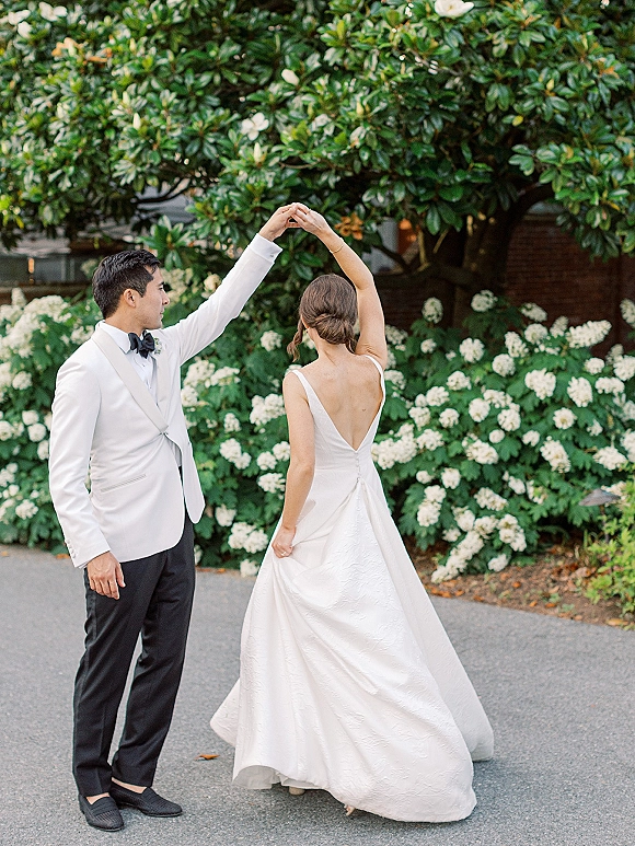 Couple portrait of groom spinning bride twirling dress in an open-back white gown on a driveway beside hydrangea shrubs and trees