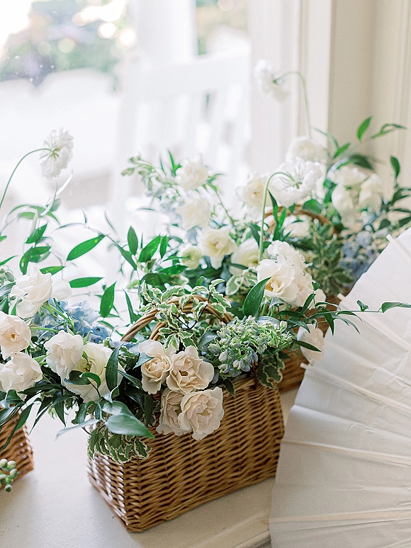 Wedding welcome baskets with white and green florals, tissue paper, and wicker baskets arranged on a white table in bright window light