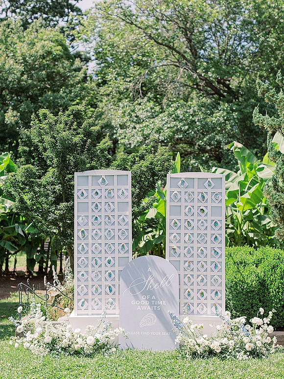 Wedding seating chart with escort card display on a white arched board, set on garden lawn with floral ground arrangement and wrought iron fence
