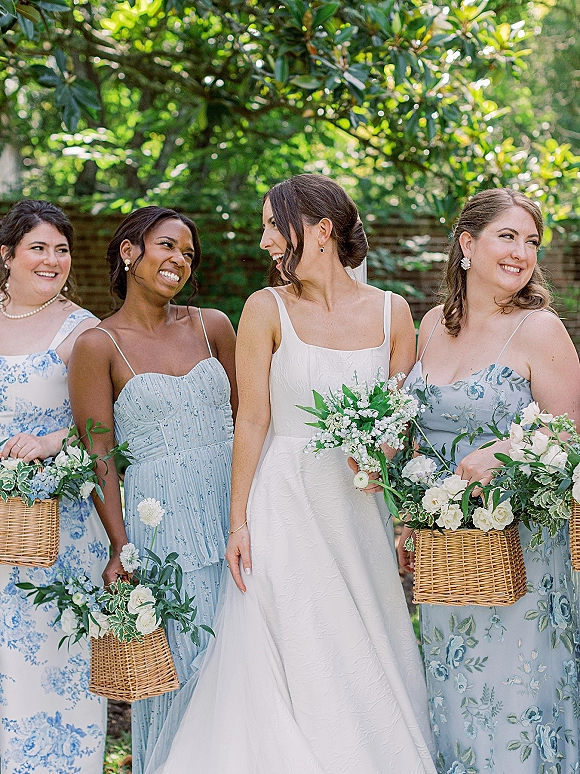 Bridesmaid group photo with bride and bridesmaids holding wicker basket bouquets, laughing on a sunny lawn by garden trees and brick wall