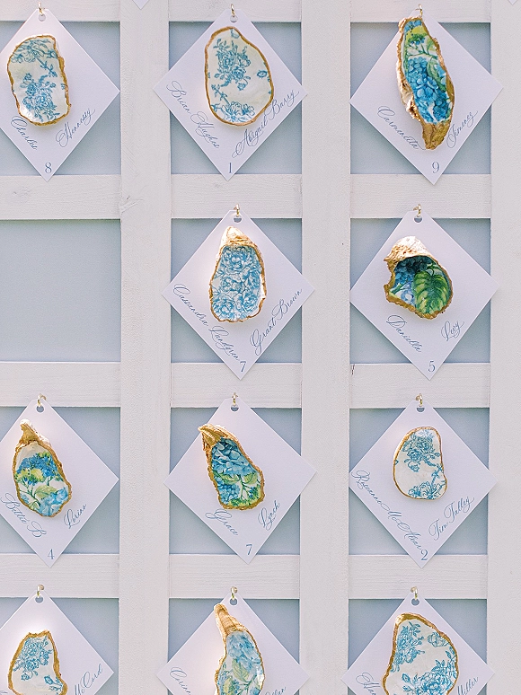 Wedding escort cards on oyster shells with hand calligraphy and gold leaf edges, hanging on hooks on a white lattice display with pale blue backing