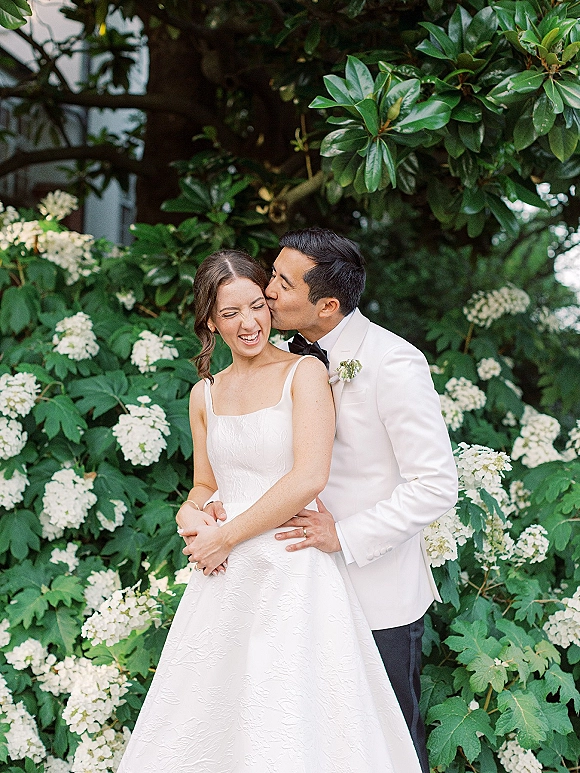 Wedding couple portrait of groom kissing bride cheek as she laughs, embracing in garden greenery with white hydrangea backdrop