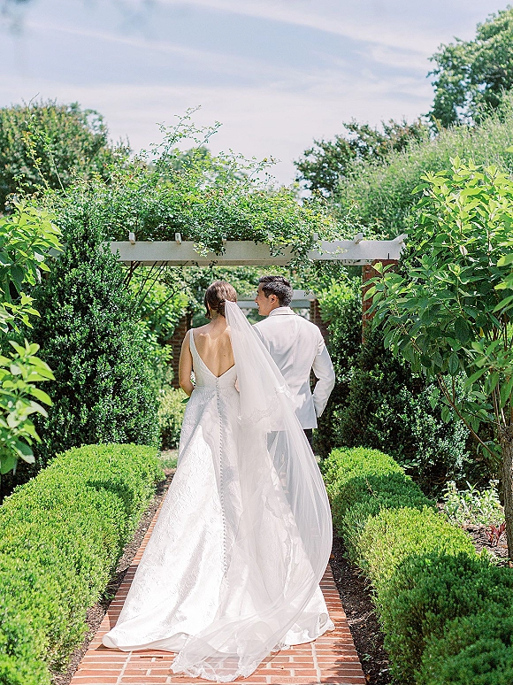 Couple portrait of bride and groom walking away, her cathedral veil flowing behind on a brick walkway through a garden pergola.