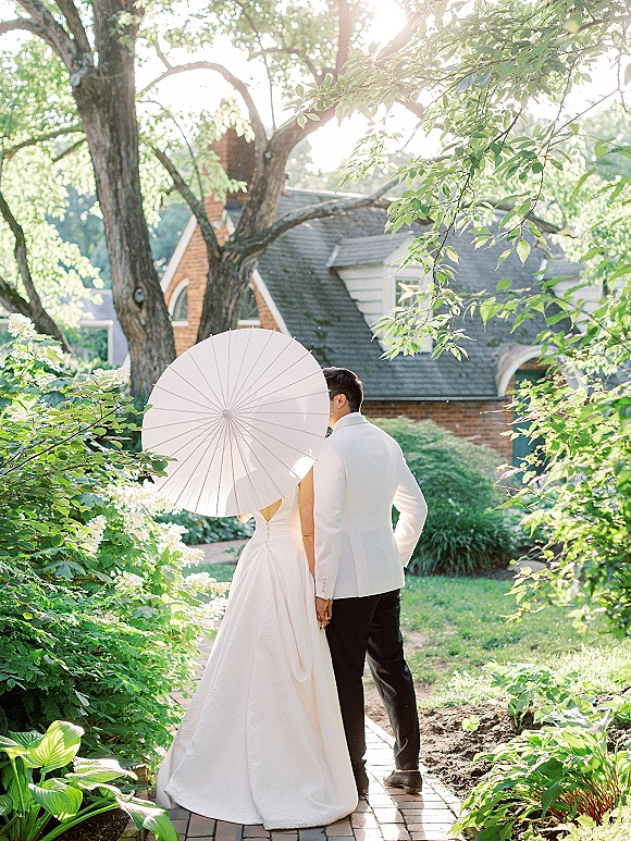 Couple portrait of bride and groom walking away hand in hand, bride in ball gown holding a parasol on a brick garden path under trees