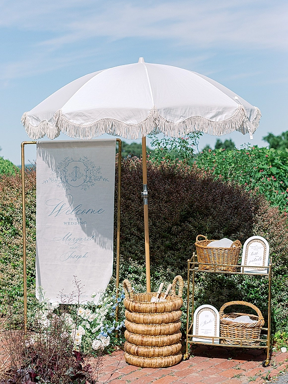 Wedding welcome sign on a gold stand with a white fringe parasol, wicker baskets for programs and towels, and florals on a brick walkway