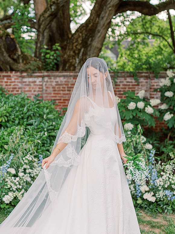 Bridal portrait of a bride with veil, looking down in a textured satin gown, holding a green-and-white bouquet by a brick garden wall