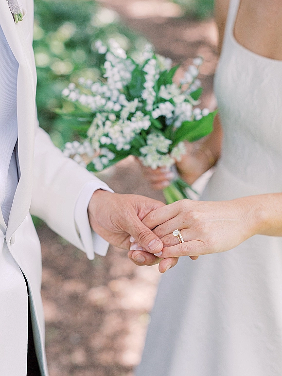 Couple holding hands, wedding hand detail highlighting engagement ring and bands, with lily of the valley bouquet accent in sunlit greenery background