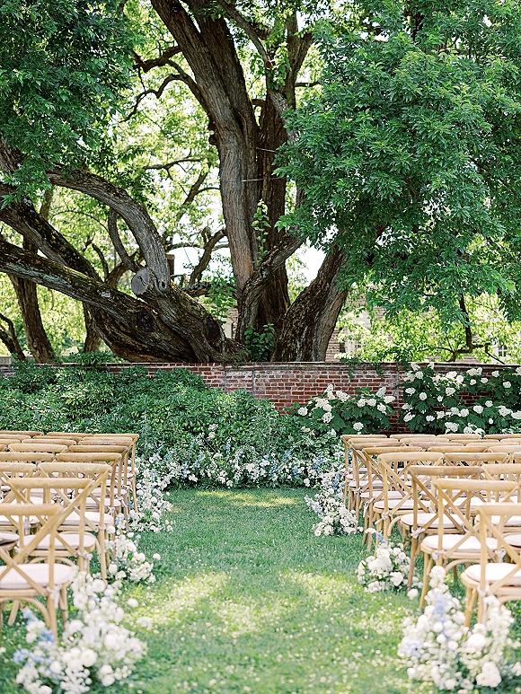 Ceremony aisle decor with outdoor wedding ceremony setup of wood cross-back chairs and white-blue ground florals on a lawn under an oak tree