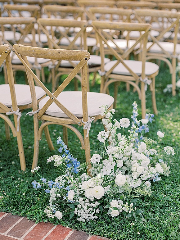 Ceremony seating with cross back chairs wedding style, cushioned rows with tied bows beside white and blue aisle florals on lawn path