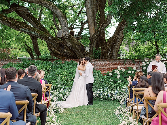 Wedding kiss as bride in a long veil and groom in a white dinner jacket embrace on a garden aisle lined with white and blue flowers