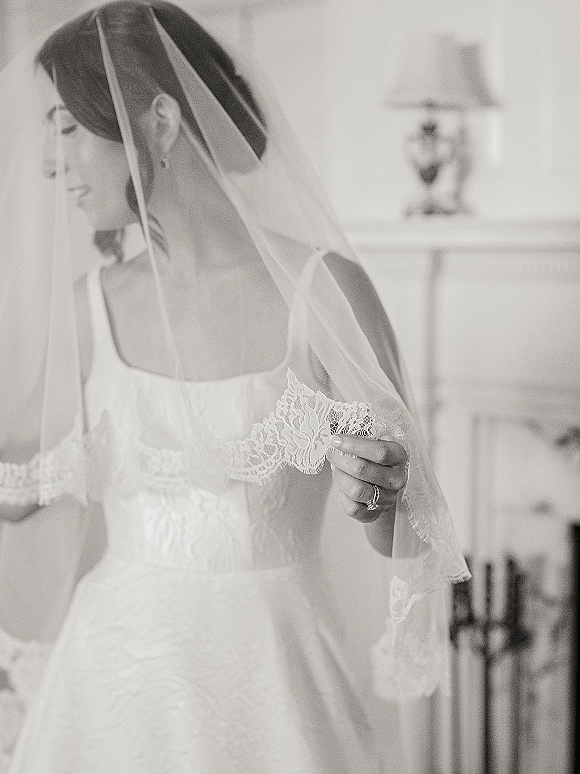 Bridal portrait of a bride in veil wearing a strapless wedding dress with lace trim, holding her veil in an indoor room by a fireplace mantel