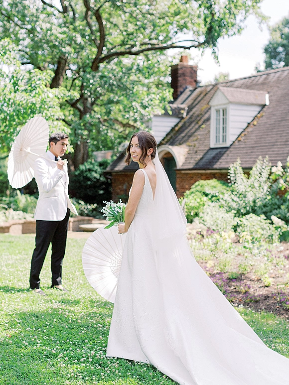 Couple portrait in a garden wedding portrait, bride in veil and open-back dress holding bouquet beside groom with parasol on lawn by cottage