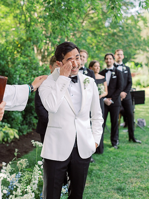 Groom crying, wiping tears in a white tuxedo jacket and black bow tie during a garden ceremony with wedding party behind