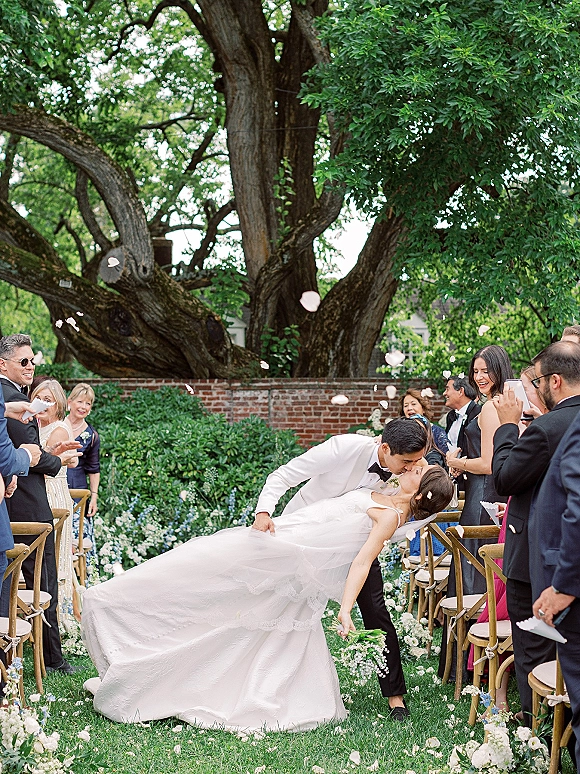 Wedding kiss portrait of couple dip kiss as guests cheer and toss flower petals, bride in ball gown holding bouquet on garden lawn aisle