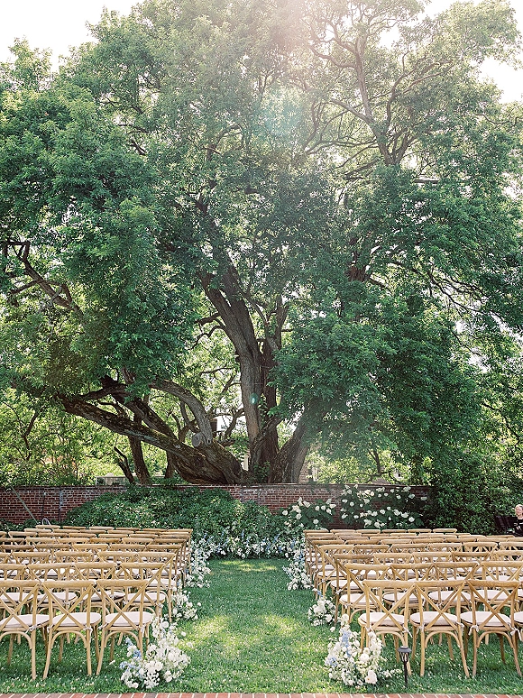 Outdoor ceremony setup with garden wedding ceremony seating, wood crossback chairs and white floral aisle markers beneath a large oak tree on grass
