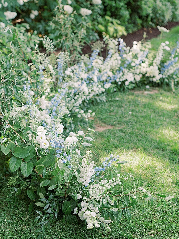Ceremony aisle florals with low white and blue blooms and greenery accents arranged along a grass lawn beside garden shrubs