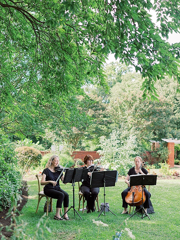Wedding musicians perform on violin and cello with sheet music stands, seated in black on a garden lawn beside a brick wall
