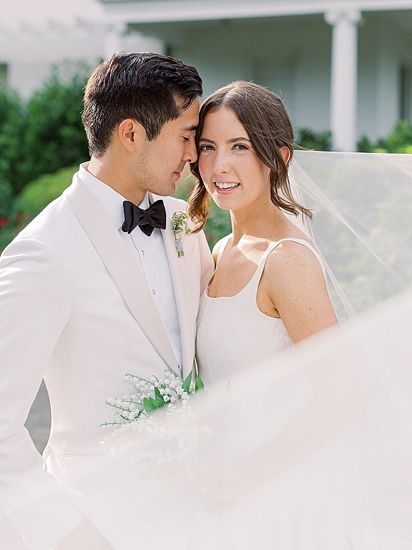 Couple portrait of groom kissing bride’s forehead as she smiles, veil and lily of the valley bouquet on a columned porch with greenery