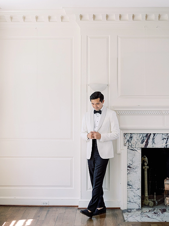 Groom portrait in a white tuxedo jacket with black bow tie standing by a marble fireplace and paneled wall, hands clasped on hardwood floor