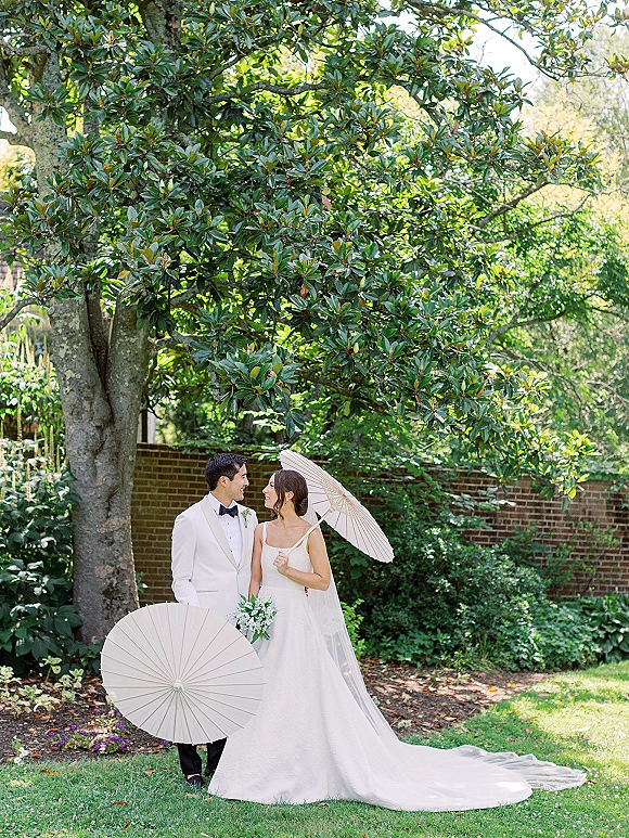 Couple portrait of bride and groom smiling, she holds a bouquet, both with white parasols on a garden lawn by a brick wall