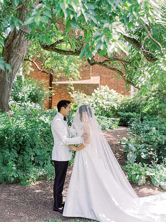 Couple portrait of bride and groom holding hands, bride in long veil and bouquet, groom in white tuxedo on a shaded garden path by brick building