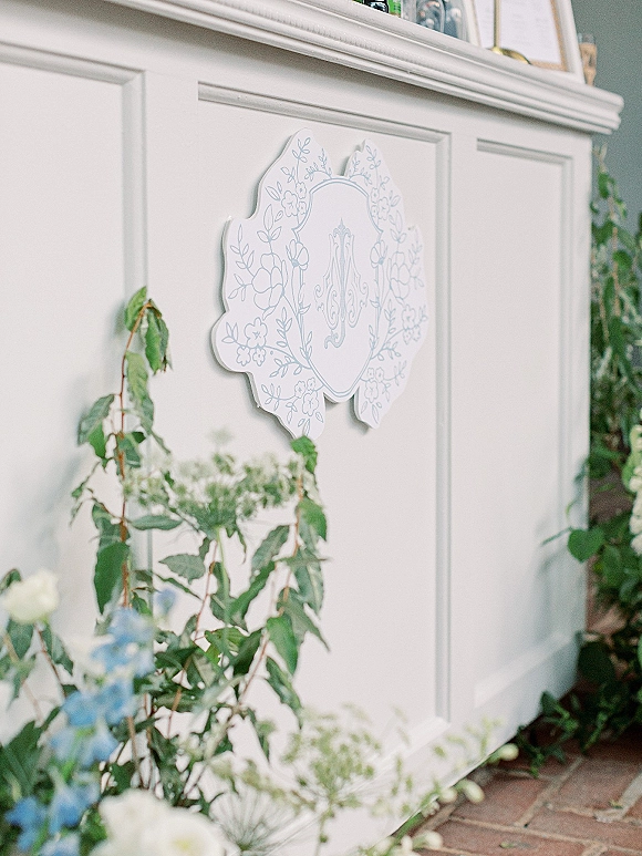 Wedding monogram sign with custom wedding monogram, scalloped edge and floral illustration framed by ivy garland and blue blooms on a white bar front
