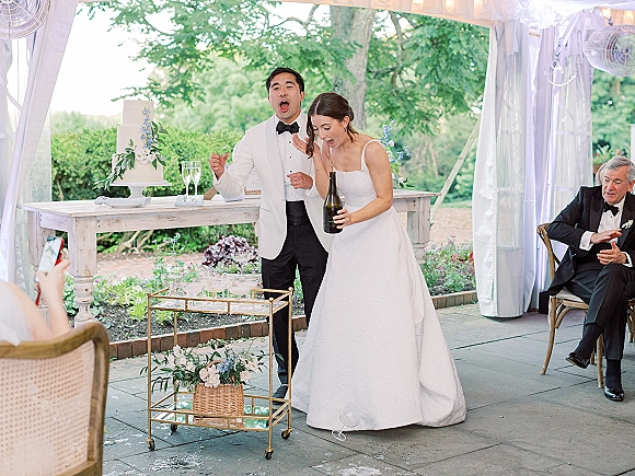 Champagne pop moment with champagne spray celebration as bride and groom laugh by a bar cart under string lights in a garden tent reception