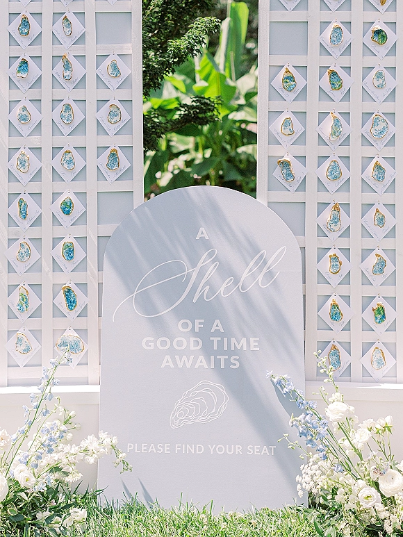 Wedding seating chart with calligraphy escort card display, oyster shell place cards on white lattice, framed by blue and white florals in sunlit garden
