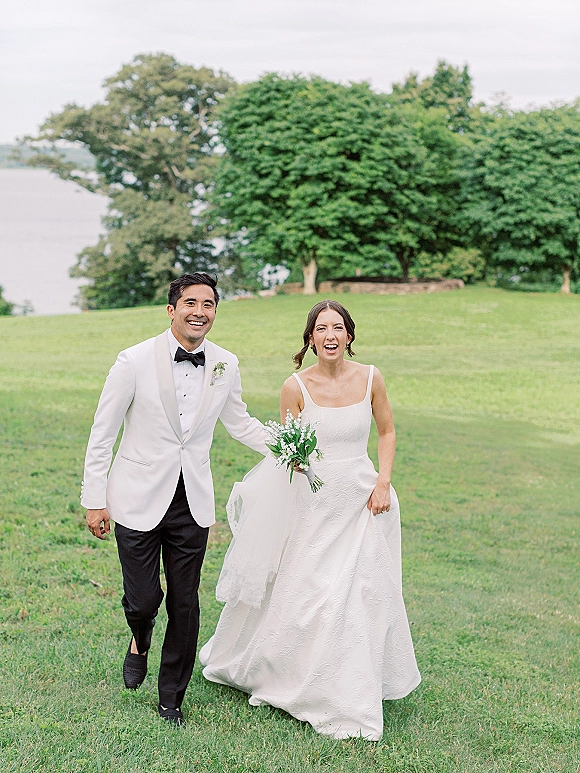 Couple portrait of bride and groom walking hand in hand, her bouquet and his white dinner jacket beside a lakeside lawn with trees