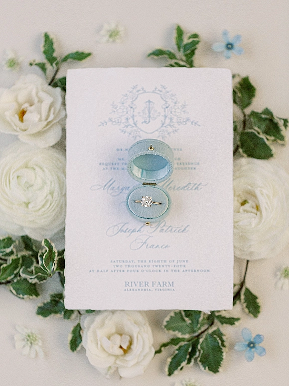 Engagement ring flatlay featuring a diamond solitaire in a blue velvet ring box atop a script wedding invitation with white flowers and greenery