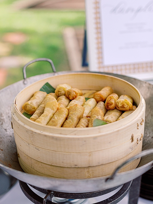 Wedding appetizers arranged in a bamboo steamer with crisp spring rolls on a banana leaf liner, set on an outdoor lawn near a blurred sign
