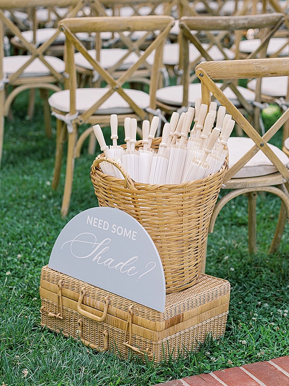 Wedding ceremony fans arranged as white hand fans in a wicker basket beside a suitcase and sign by crossback chairs on a lawn walkway