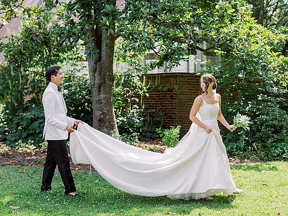 Couple portrait of bride and groom outdoors, groom holding her dress train as she looks back with a white bouquet on a garden lawn by brick building