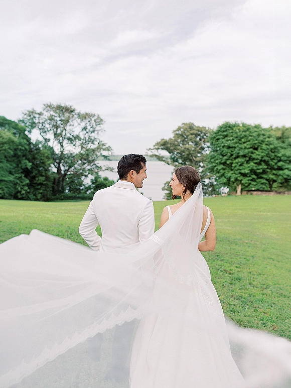 Couple portrait of bride and groom walking away on a green lawn, bride glancing back as her long veil trails under cloudy sky