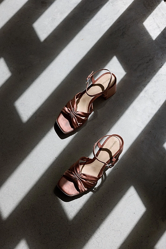 Bridal shoes flatlay showing blush satin ankle strap block heels on a concrete floor, with window light shadows highlighting the texture