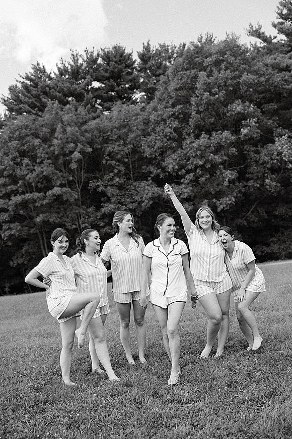 Bridesmaids getting ready in bridesmaid pajama sets, laughing together in matching button-up tops and shorts in a grassy field