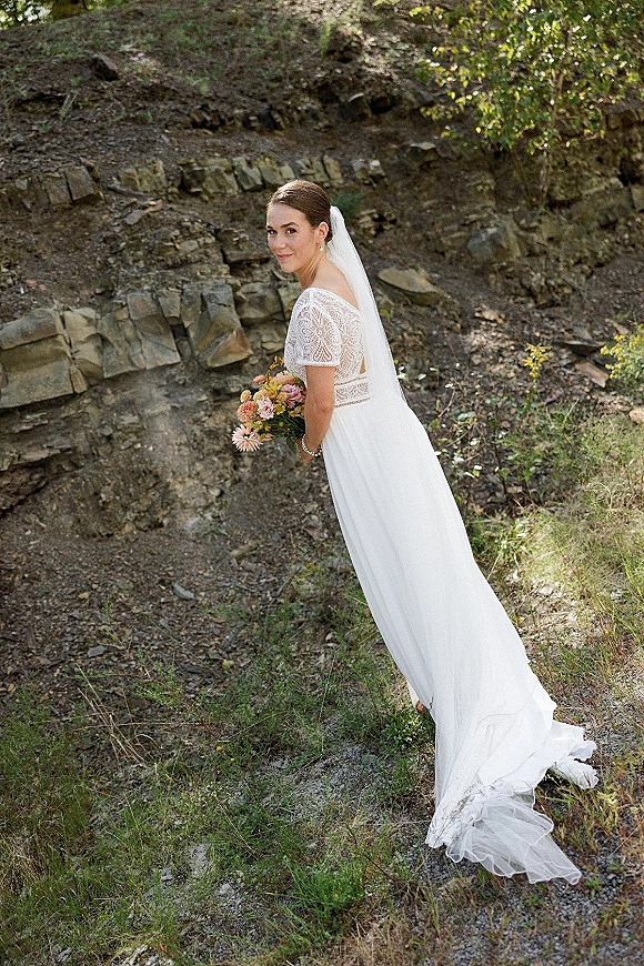 Bridal portrait of a bride holding bouquet in a lace wedding dress and veil, looking back on a rocky hillside with trees and grass