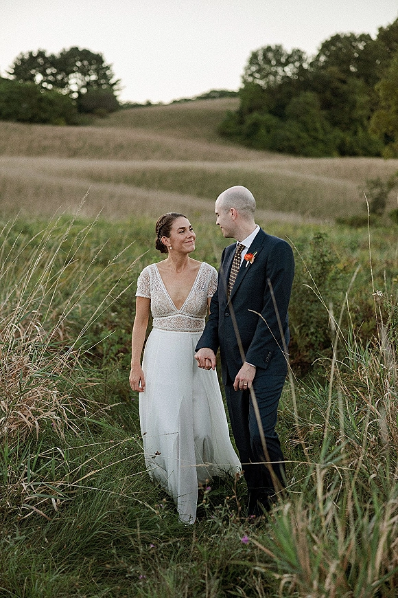 Couple portrait of bride and groom holding hands in tall grass, her lace bodice dress and his boutonniere under overcast hills