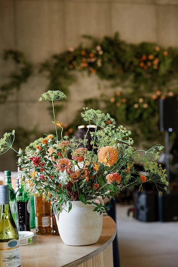 Wedding centerpiece with wildflower wedding centerpiece blooms in a ceramic vase, orange flowers and greenery on a wood table with wine bottles