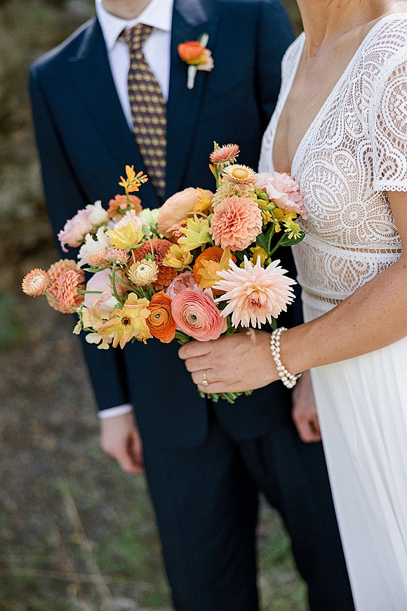 Bridal bouquet of colorful bridal bouquet blooms held by a bride in a lace dress, with pearl bracelet and ring, set against outdoor greenery