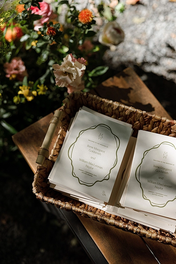 Wedding programs with ceremony program design in green ink in a wicker basket beside roses and greenery on a wooden table in sunlight
