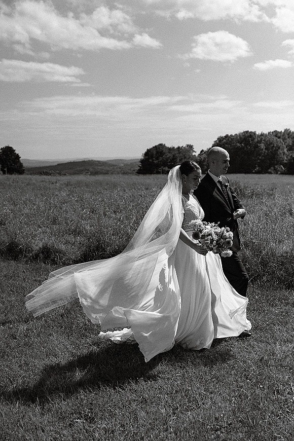Couple portrait in a black and white wedding portrait, bride holding a daisy bouquet as they walk through a meadow under cloudy hills