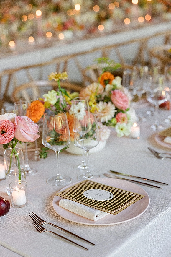 Reception tablescape with wedding place setting, floral centerpiece and bud vases, votive candles, gold cutlery on a long banquet table under string lights