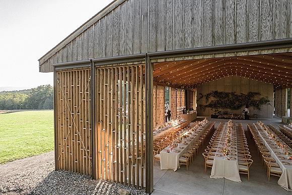 Reception tablescape with long banquet tables in white linens, taper candles and floral centerpieces under string lights in a barn