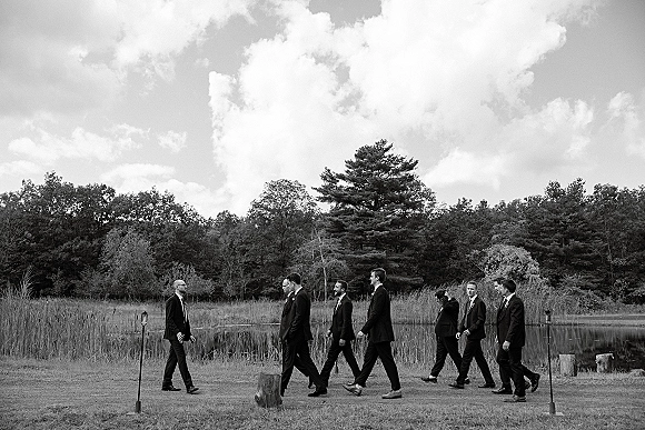 Groomsmen portrait of men in matching suits with ties and boutonnieres, walking through meadow grass by a pond under cloudy sky